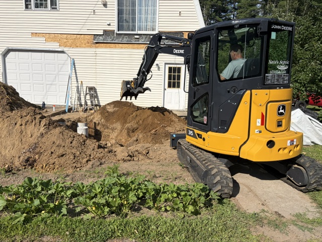 Nate's Septic branded John Deere 35 mini excavator digging a septic trench beside a home's side entry in New Ipswich, NH