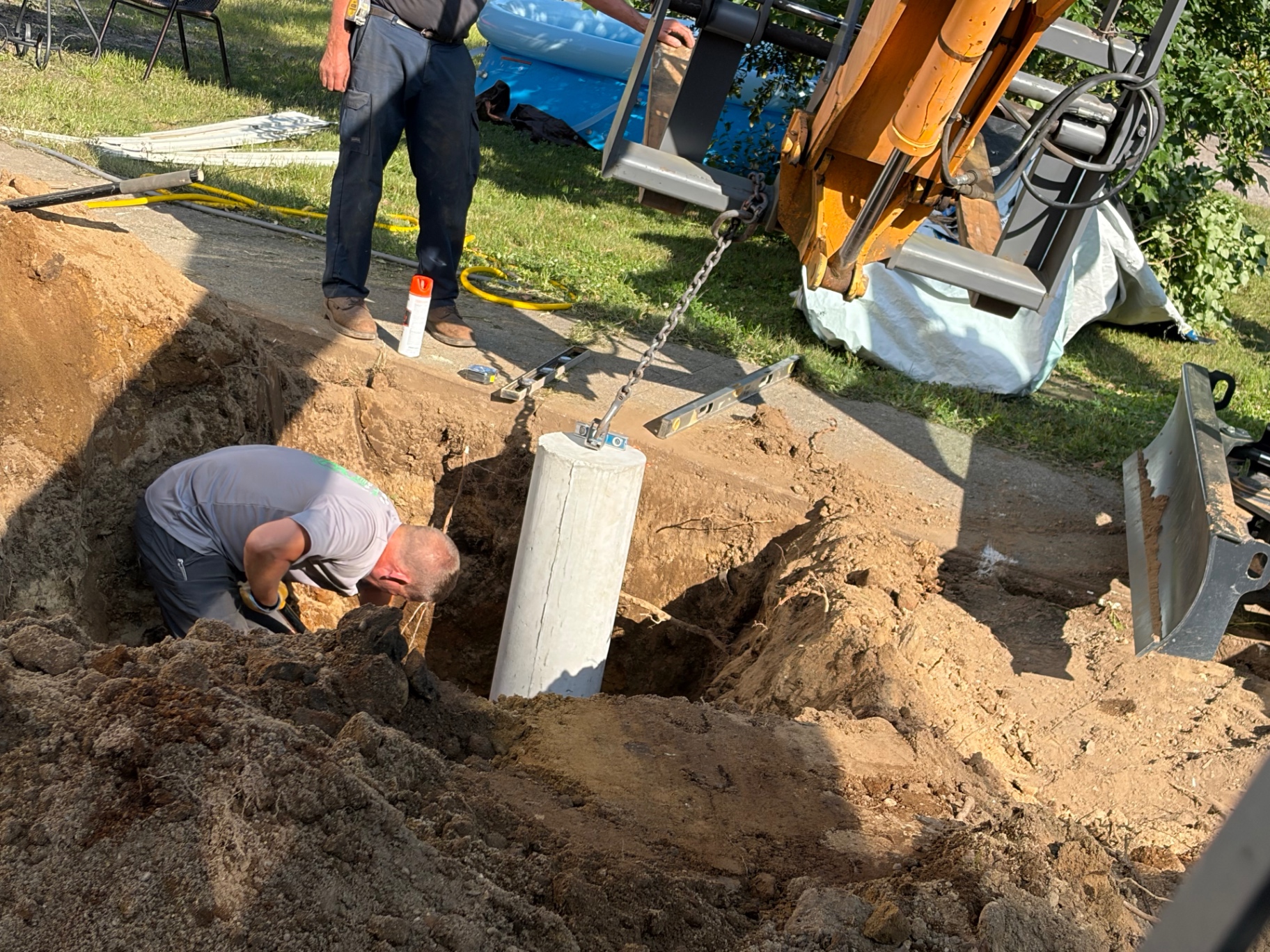 Crew setting a concrete septic riser into an excavated hole, leveled with a chain off the mini excavator